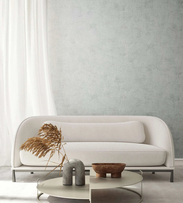 Marburg City Glow 34281 Gray mottled wallpaper forms a feature wall behind a curved white sofa, a round low coffee table with a sculptural object, a small bowl, and dried stems, with a sheer curtain at the left.