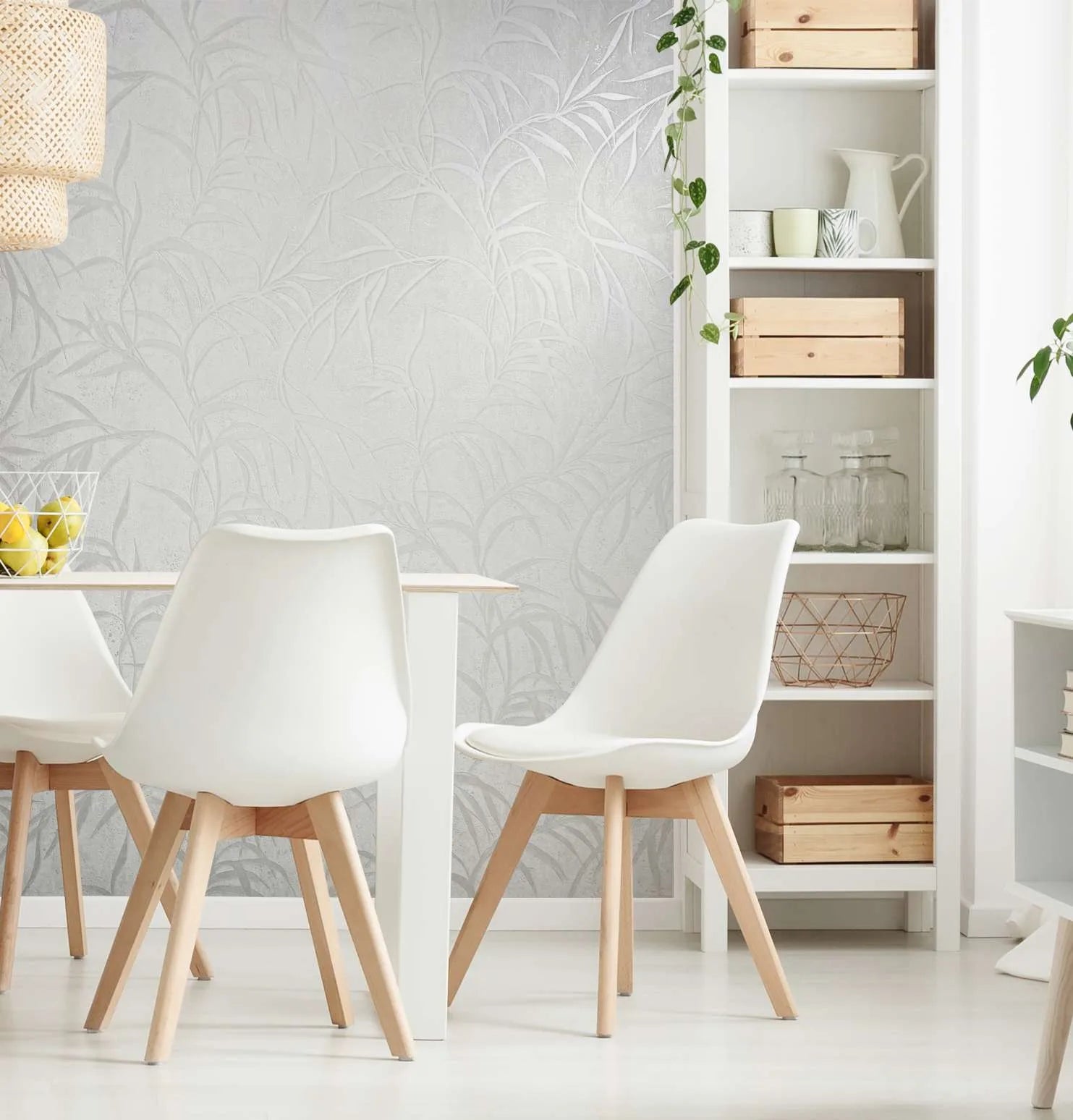 Marburg City Glow 34283 Light gray-white wallpaper with a tonal leafy leaf pattern behind a modern dining area with white chairs, a white table, open shelving, and a woven pendant; a wire bowl of lemons sits on the table.