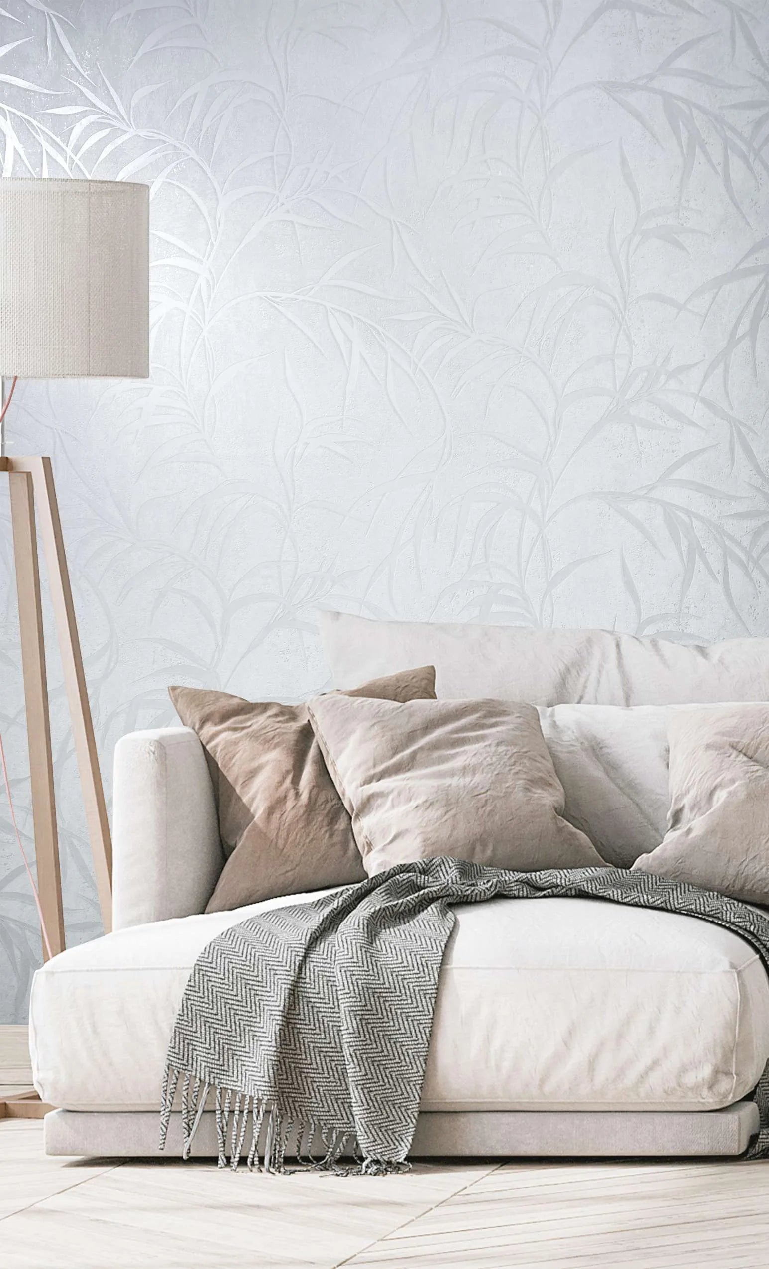 Marburg City Glow 34283 Tonal white and light gray wallpaper with overlapping leaf fronds behind a low sofa with neutral pillows and a wooden tripod floor lamp.