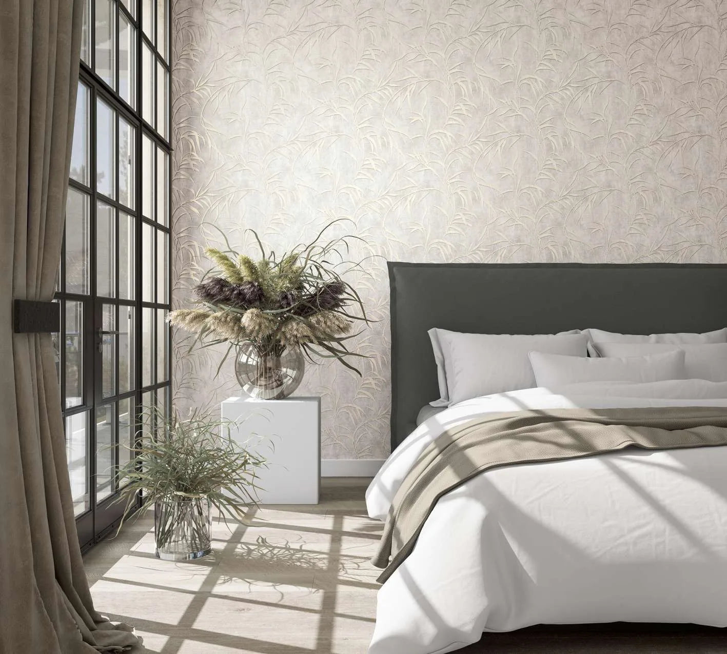 Marburg City Glow 34284 Light beige wallpaper with a subtle grass‑leaf pattern behind a dark upholstered headboard in a sunlit bedroom, next to large grid windows with curtains and glass vases filled with grasses.