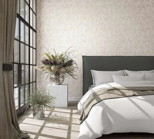 Marburg City Glow 34284 Light beige wallpaper with a subtle grass‑leaf pattern behind a dark upholstered headboard in a sunlit bedroom, next to large grid windows with curtains and glass vases filled with grasses.