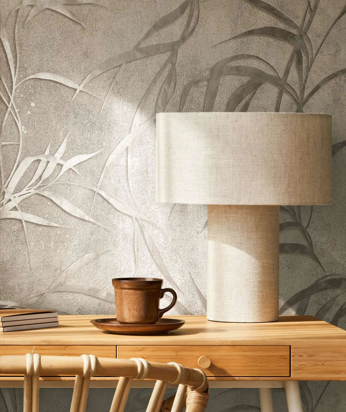 Marburg City Glow 34286 Gray leaf-pattern wallpaper behind a light fabric table lamp on a wooden desk with a brown cup and saucer, a small stack of books, and part of a wooden chair.