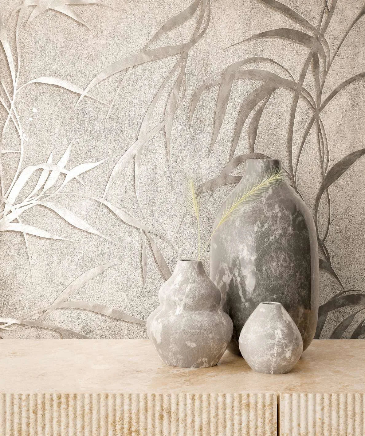 Marburg City Glow 34286 Tonal gray wallpaper with large slender leaf motifs is shown behind three stone-look vases on a light textured console.