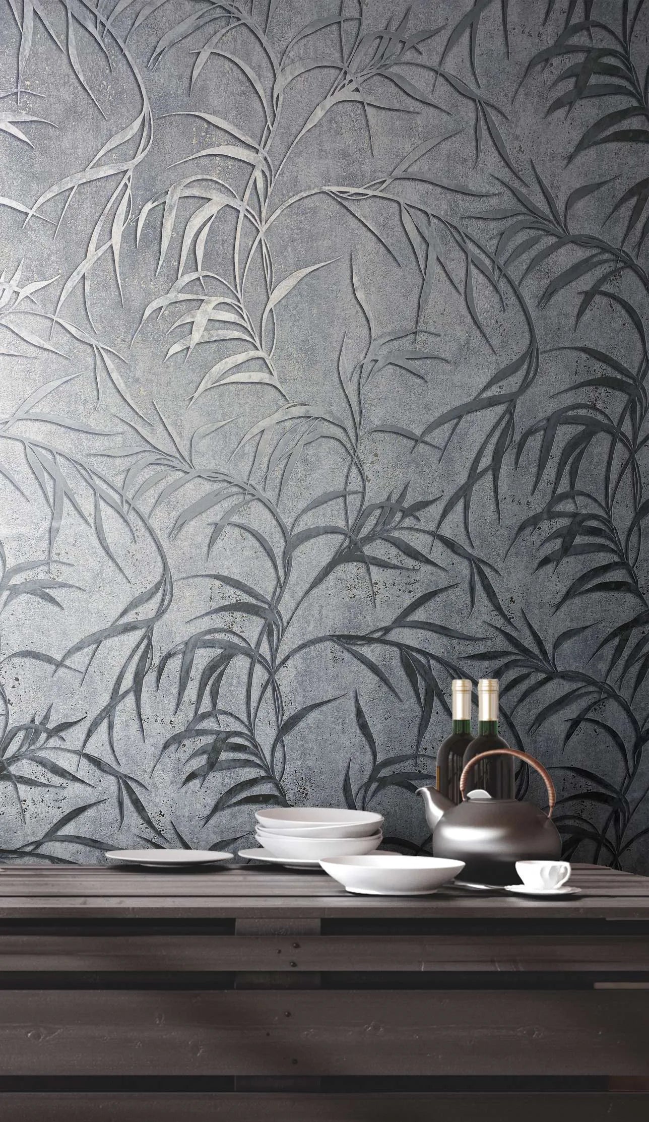 Marburg City Glow 34287 Gray wallpaper with a large leafy botanical design shown behind a dark wood table set with stacked white plates, bowls, a teapot, a cup and saucer, and two bottles.