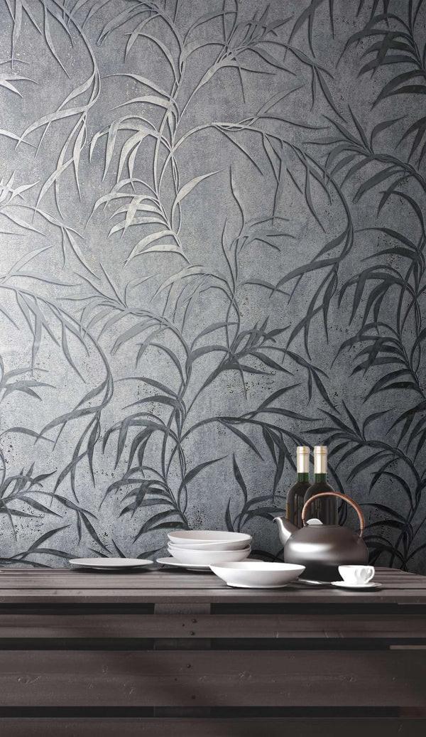 Marburg City Glow 34287 Gray wallpaper with a large leafy botanical design shown behind a dark wood table set with stacked white plates, bowls, a teapot, a cup and saucer, and two bottles.