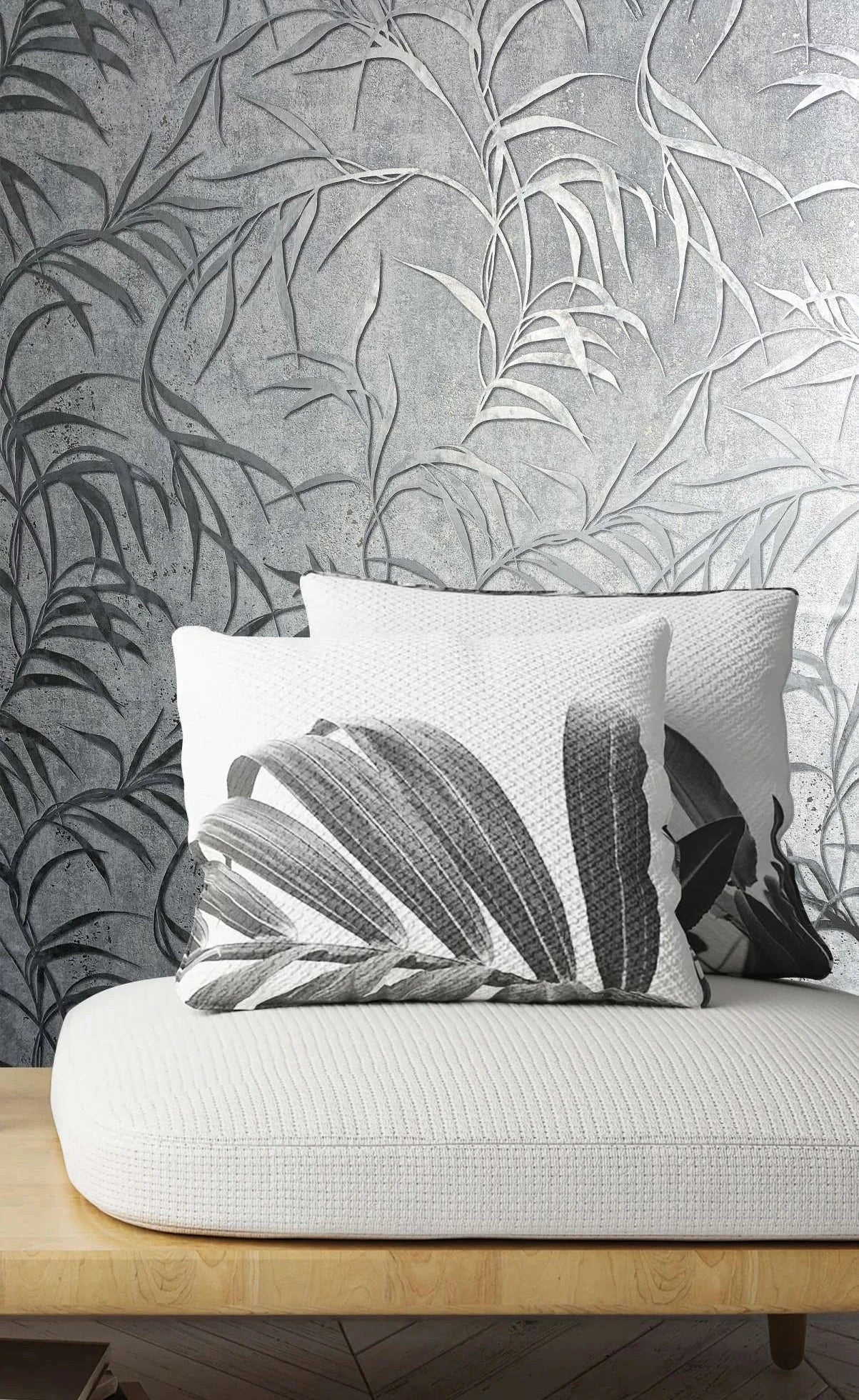 Marburg City Glow 34287 Gray wallpaper with large leafy fronds on a textured background behind a light wood bench with a white cushion, two monochrome botanical pillows, a herringbone floor, and a small stack of books.