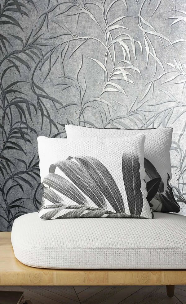 Marburg City Glow 34287 Gray wallpaper with large leafy fronds on a textured background behind a light wood bench with a white cushion, two monochrome botanical pillows, a herringbone floor, and a small stack of books.