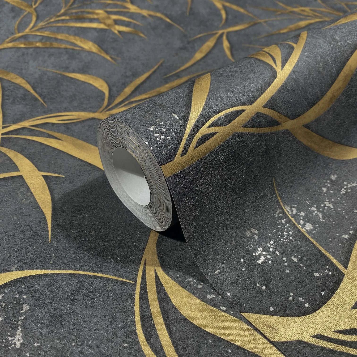 Marburg City Glow 34289 Close-up of a wallpaper roll partially unrolled, showing a gold botanical branch pattern on a dark charcoal textured background with light speckled accents.