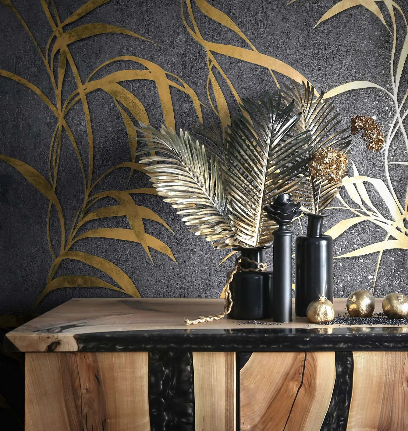Marburg City Glow 34289 Dark black wallpaper with large gold botanical fronds behind a wood console styled with black vases, decorative gold leaves, and small gold orbs.
