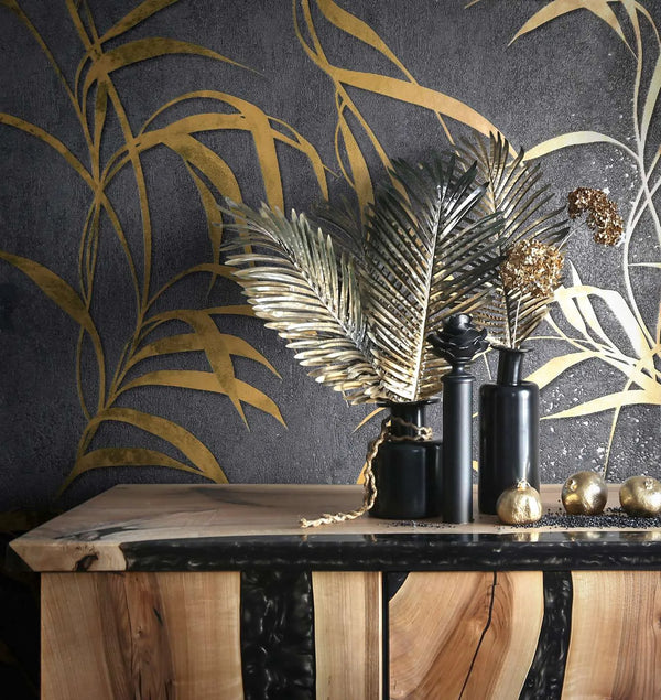 Marburg City Glow 34289 Dark black wallpaper with large gold botanical fronds behind a wood console styled with black vases, decorative gold leaves, and small gold orbs.