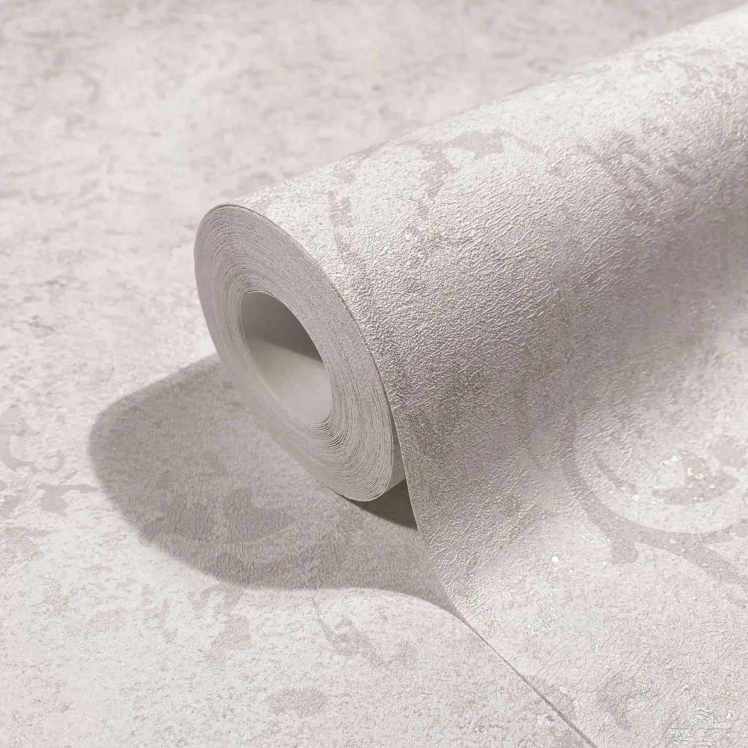 Marburg City Glow 34290 Close-up of a wallpaper roll resting on matching surface, showing light-toned textured wallpaper with a subtle tonal scroll pattern and soft sheen.