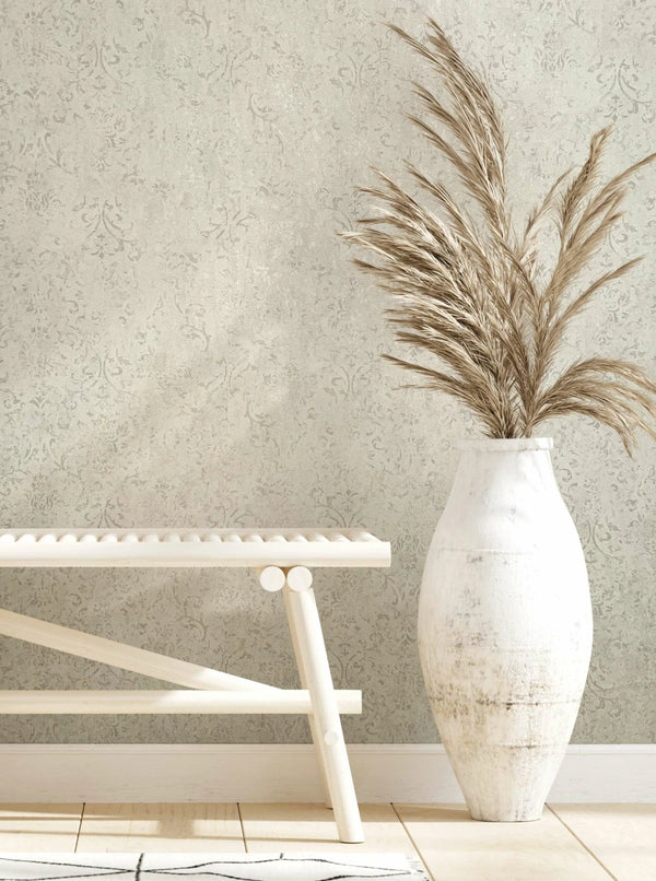 Marburg City Glow 34290 Light neutral wallpaper with a subtle tonal damask pattern shown in a styled scene with a pale bench, a tall white ceramic vase holding pampas grass, baseboard, light wood floor, and a small edge of a rug.