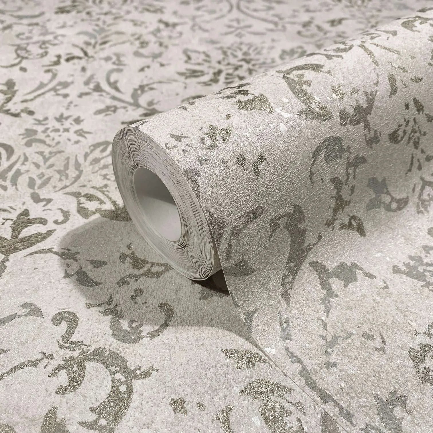 Marburg City Glow 34291 Close-up of a wallpaper roll with a textured ornamental scroll pattern in light beige and silver-gray, resting on the same patterned surface.