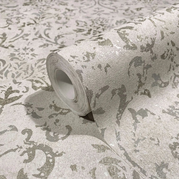 Marburg City Glow 34291 Close-up of a wallpaper roll with a textured ornamental scroll pattern in light beige and silver-gray, resting on the same patterned surface.