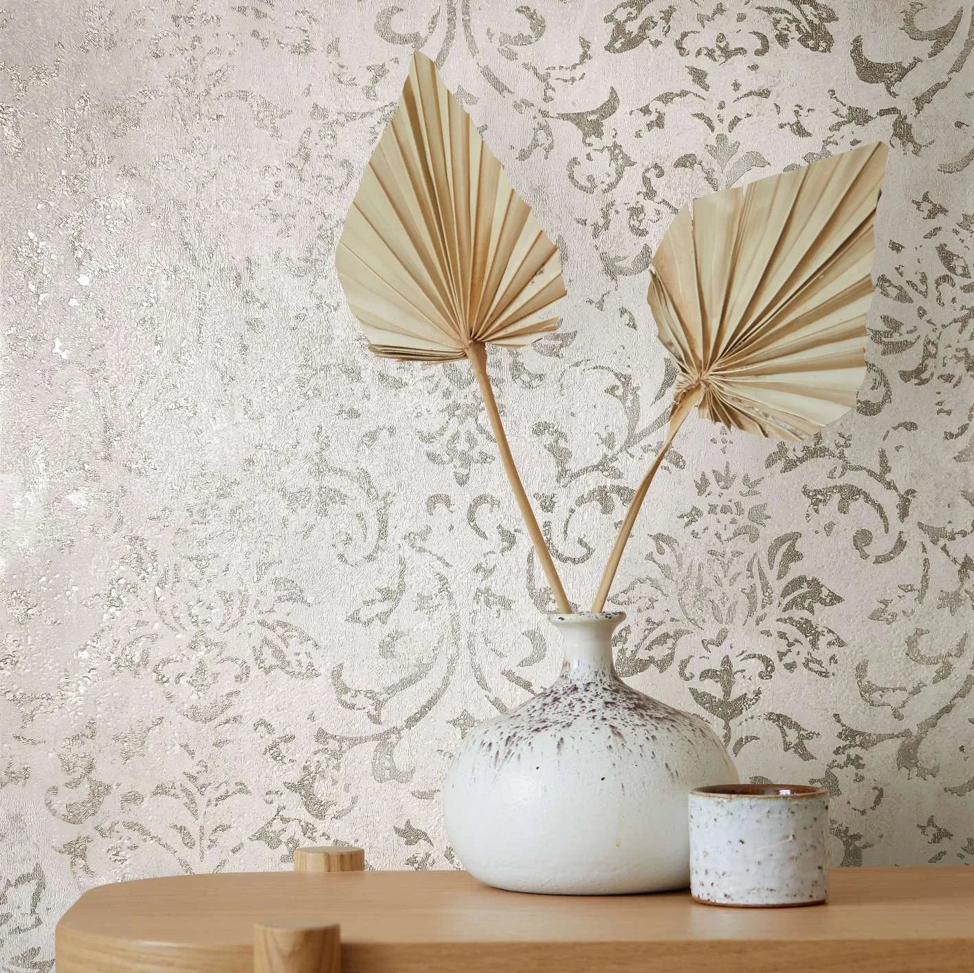 Marburg City Glow 34291 Styled close-up of a light neutral damask wallpaper with a soft sheen, shown behind a speckled ceramic vase holding two dried palm leaves and a small ceramic cup on a wooden surface.