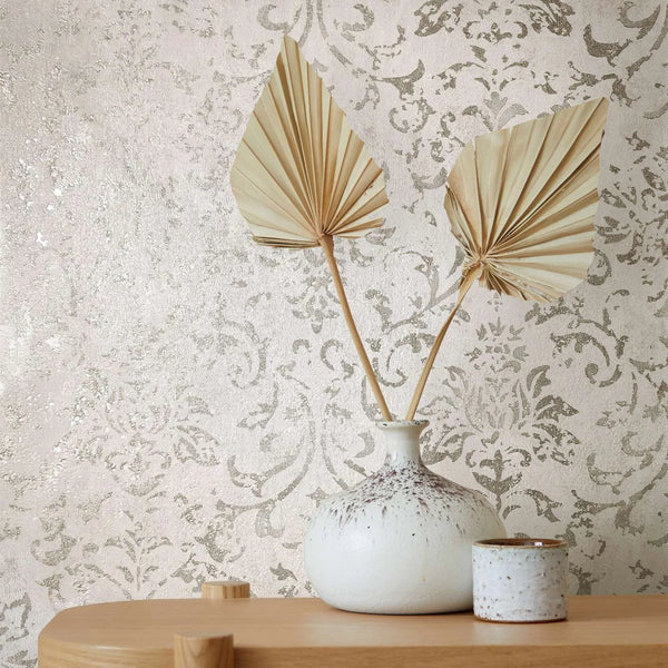 Marburg City Glow 34291 Styled close-up of a light neutral damask wallpaper with a soft sheen, shown behind a speckled ceramic vase holding two dried palm leaves and a small ceramic cup on a wooden surface.