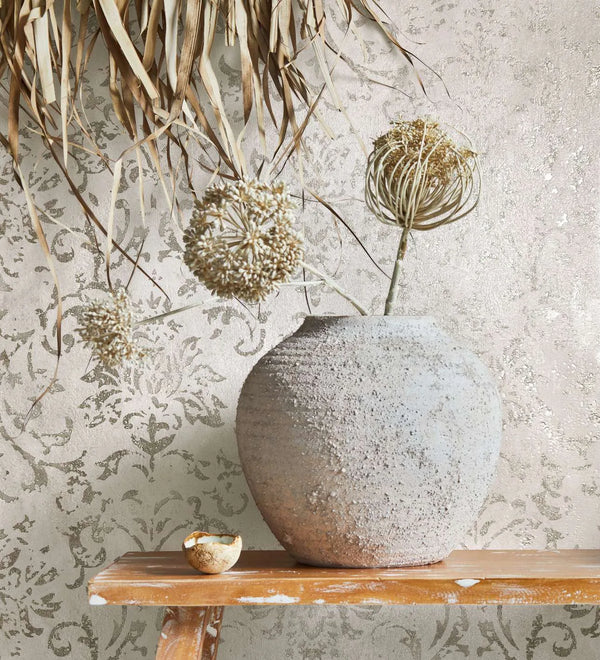 Marburg City Glow 34291 Close-up of beige damask wallpaper with a subtle shimmer behind a textured stone vase holding dried stems on a worn wooden bench, with hanging dried leaves above and a small bowl beside the vase.