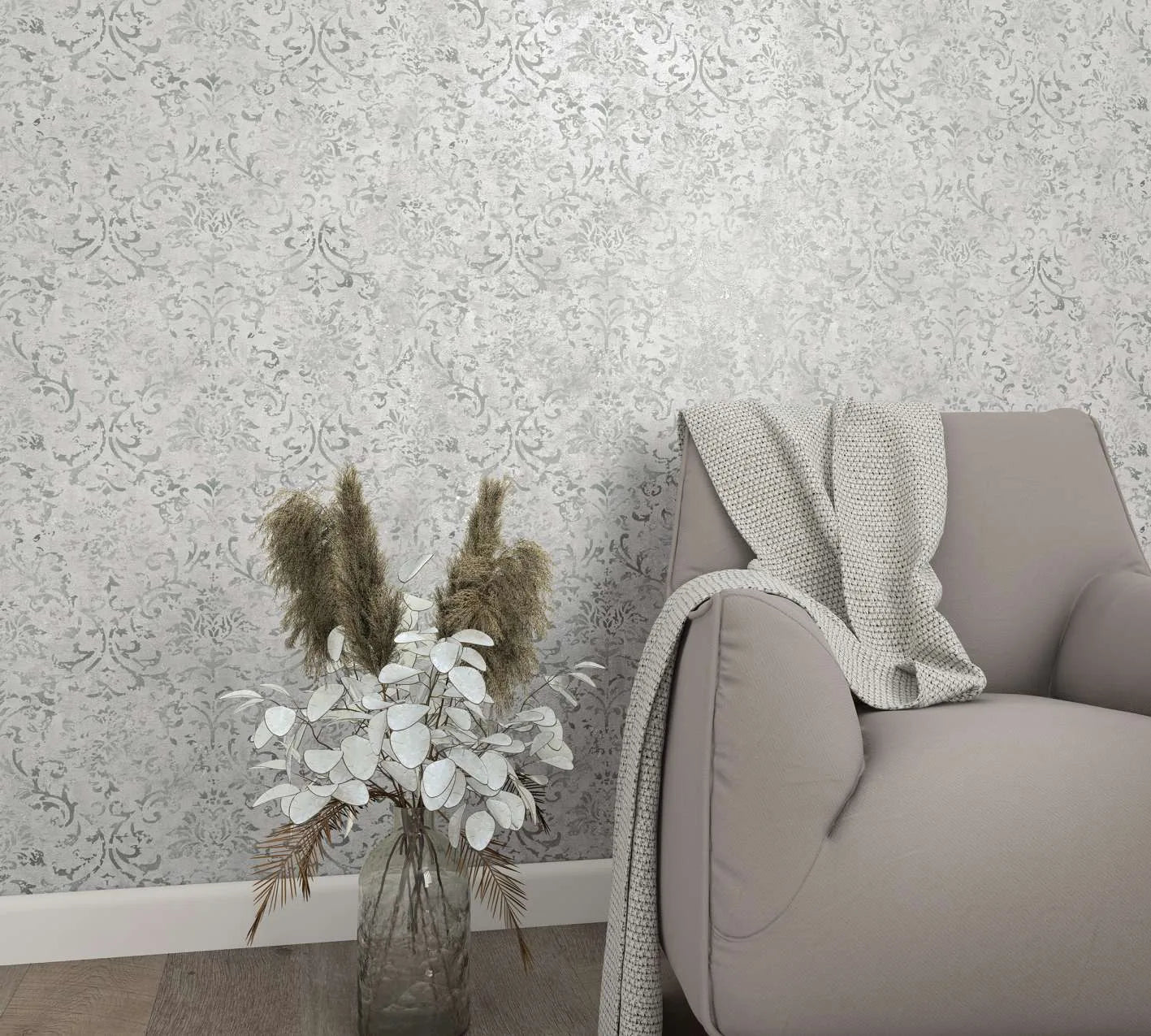 Marburg City Glow 34292 Light gray damask-patterned wallpaper shown in a living room vignette with an upholstered armchair draped with a knit throw, a glass vase of dried foliage, white baseboard, and wood flooring.