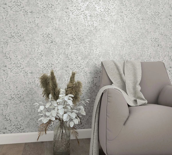 Marburg City Glow 34292 Light gray damask-patterned wallpaper shown in a living room vignette with an upholstered armchair draped with a knit throw, a glass vase of dried foliage, white baseboard, and wood flooring.