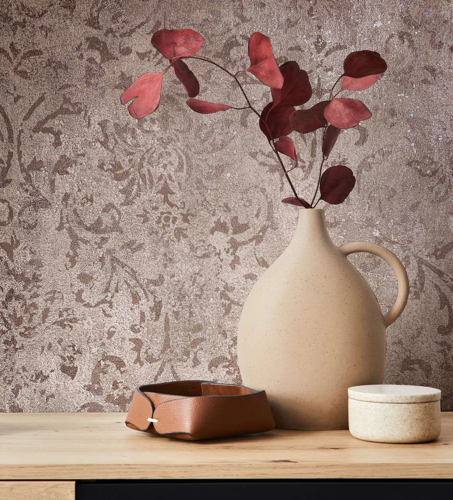 Marburg City Glow 34293 Tonal brown damask wallpaper with a textured look behind a beige ceramic jug holding red leaves, a small leather tray, and a round lidded container on a wooden surface.