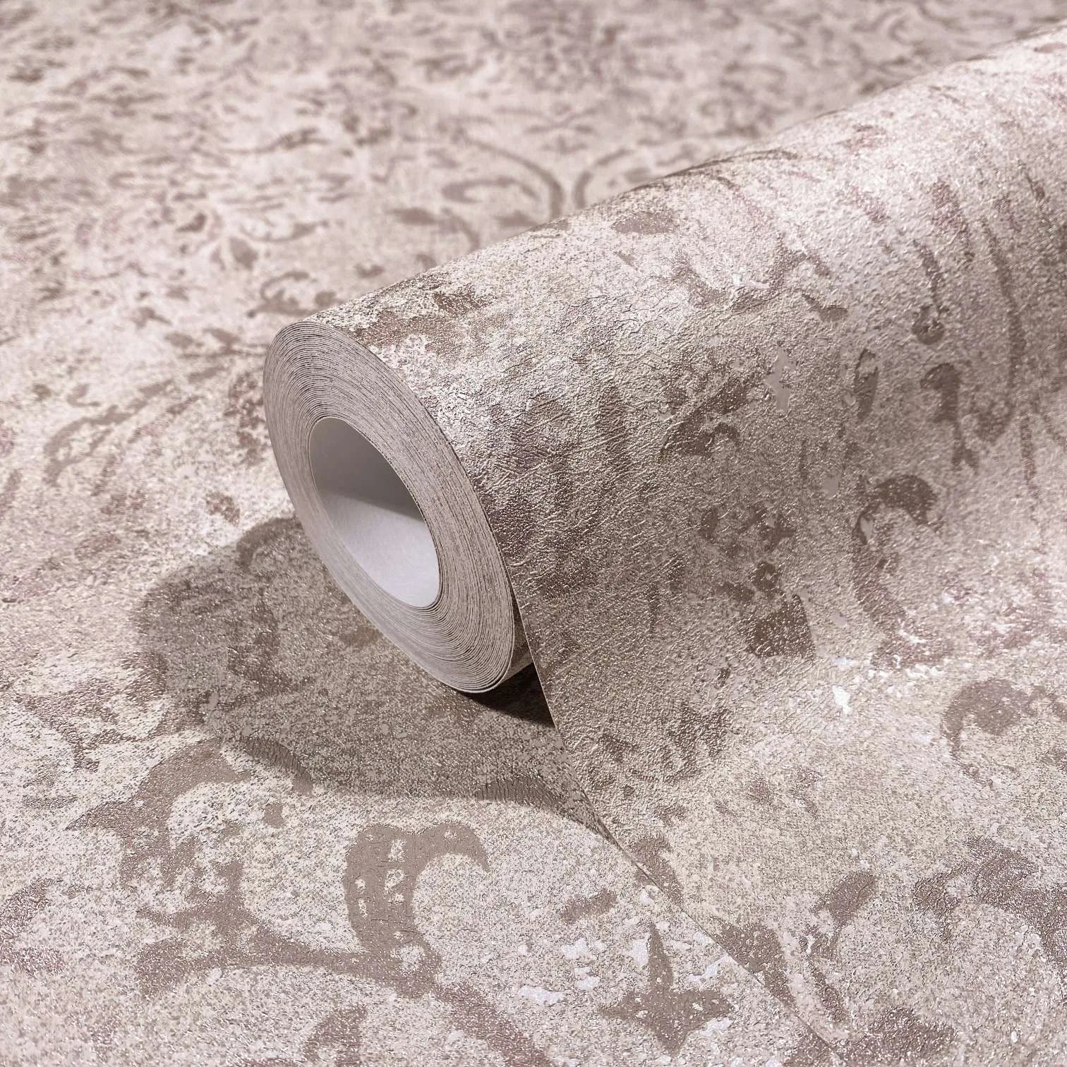 Marburg City Glow 34293 Close-up of a wallpaper roll resting on matching unrolled paper, showing a mottled textured scroll pattern in light brown tones.
