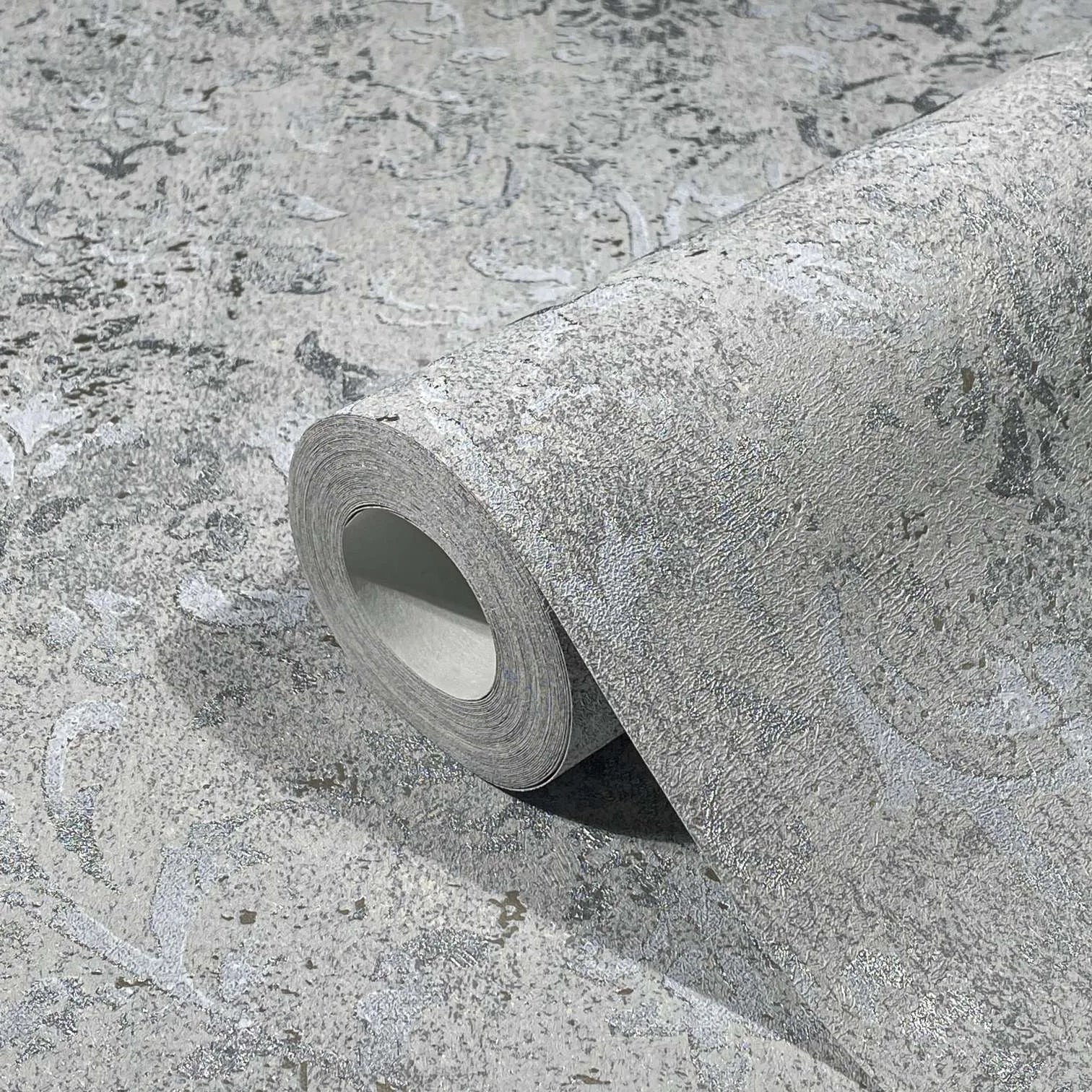 Marburg City Glow 34294 A gray textured wallpaper roll resting on the same pattern, showing a subtle scroll motif with a distressed look.