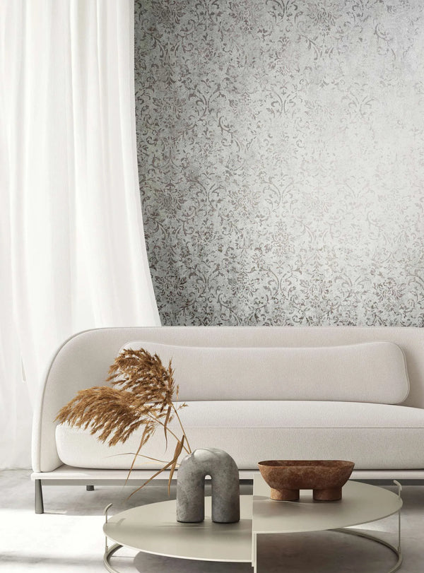 Marburg City Glow 34294 Gray wallpaper with a distressed damask pattern shown behind a light sofa, a white curtain, and a round coffee table with decor and dried grasses.