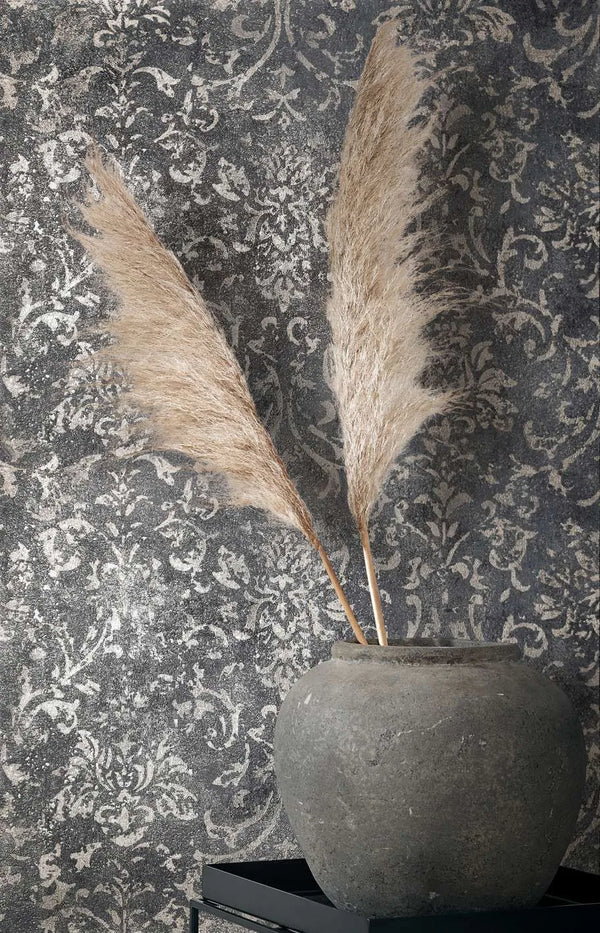 Marburg City Glow 34296 Close-up of an ornate, distressed damask wallpaper used as a backdrop, with a large stone-like pot and two pampas grass stems on a black tray in front.
