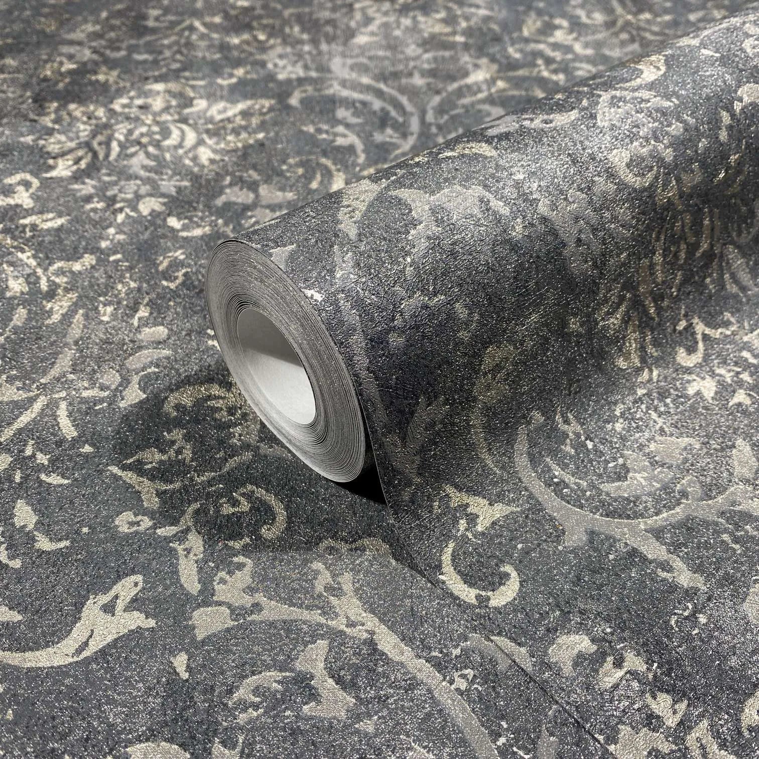 Marburg City Glow 34296 Close-up of a wallpaper roll resting on matching unrolled wallpaper, showing a charcoal and silver distressed damask-style scroll pattern with a textured look.