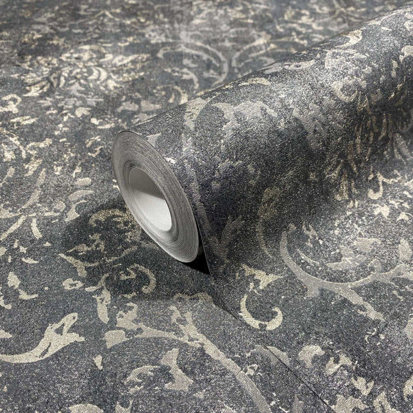 Marburg City Glow 34296 Close-up of a wallpaper roll resting on matching unrolled wallpaper, showing a charcoal and silver distressed damask-style scroll pattern with a textured look.