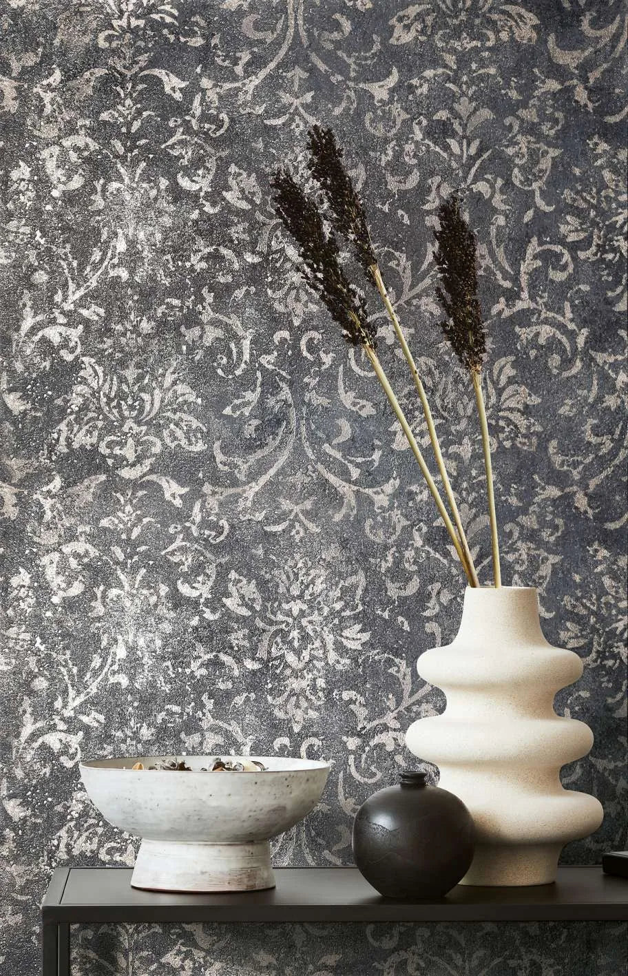 Marburg City Glow 34296 Dark charcoal wallpaper with a distressed damask pattern shown behind a sculptural light ceramic vase with tall stems, a small dark round vase, and a ceramic bowl on a narrow shelf.