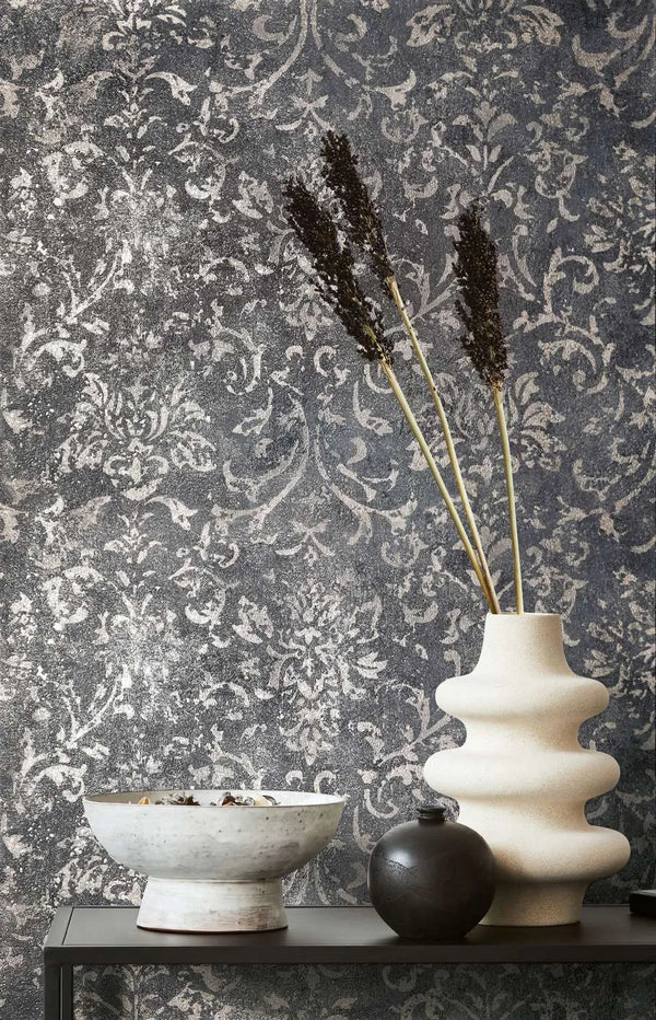 Marburg City Glow 34296 Dark charcoal wallpaper with a distressed damask pattern shown behind a sculptural light ceramic vase with tall stems, a small dark round vase, and a ceramic bowl on a narrow shelf.