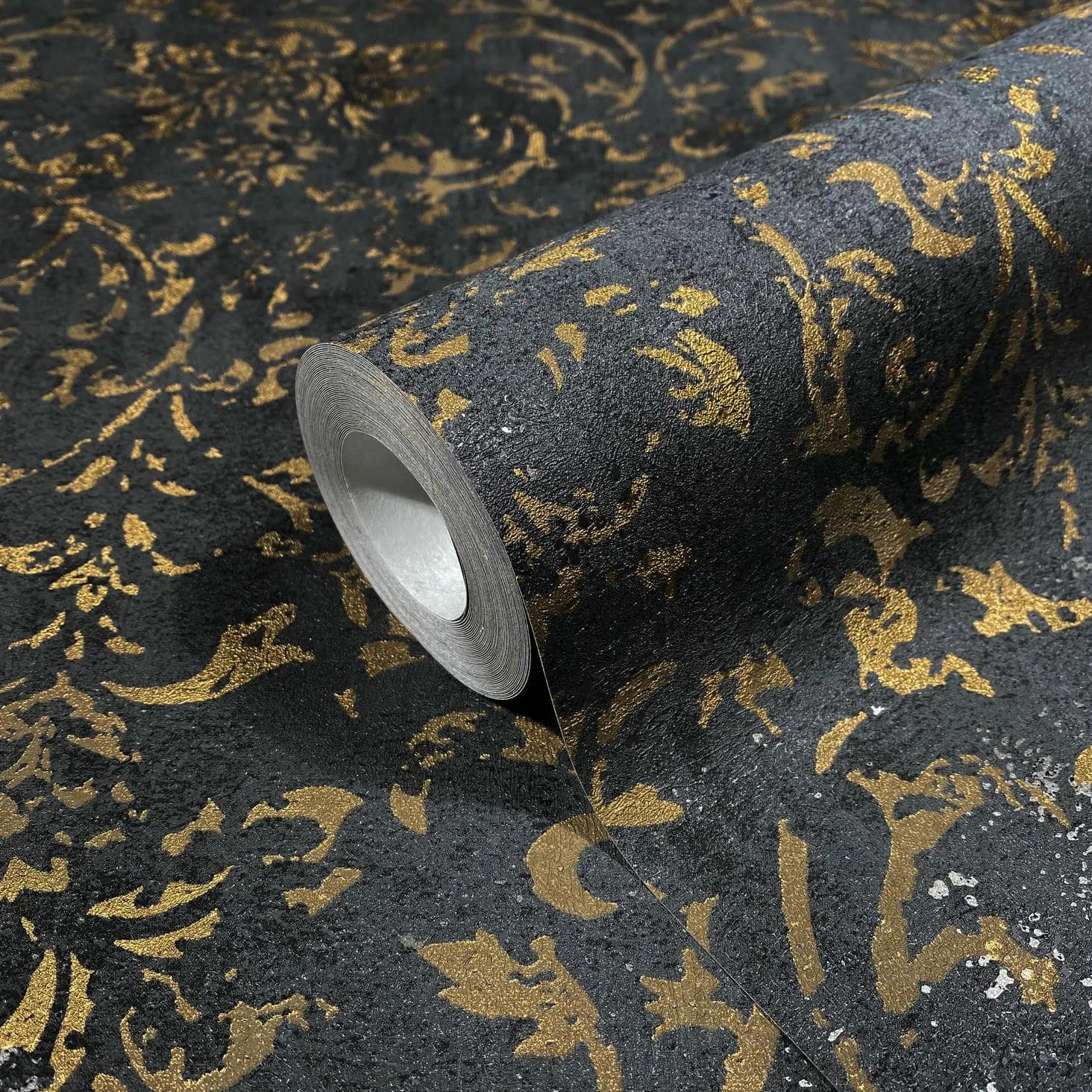 Marburg City Glow 34297 Close-up of a wallpaper roll with a metallic gold damask pattern on a dark textured background, shown partially unrolled.