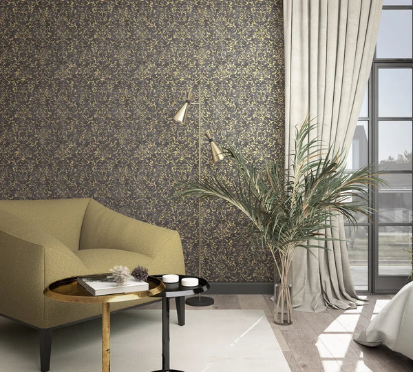 Marburg City Glow 34297 Living room scene with dark wallpaper covered in gold scrolling floral vines behind an armchair, two round side tables, a floor lamp, a tall plant in a glass vase, and a curtained window.