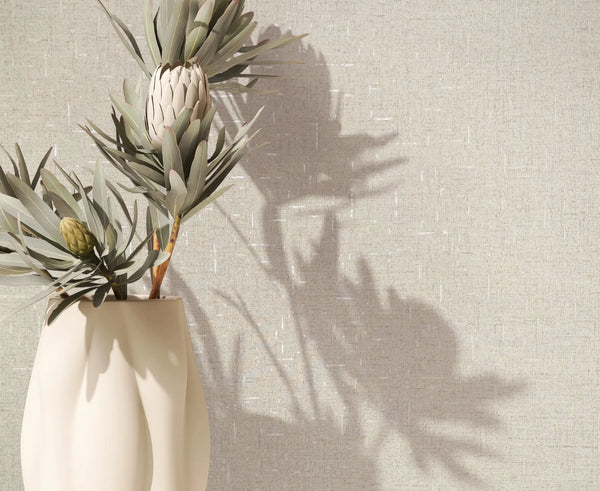 Marburg City Romance 35107 Beige linen-look textured wallpaper shown behind a cream vase holding protea stems, with plant shadows cast on the wall.