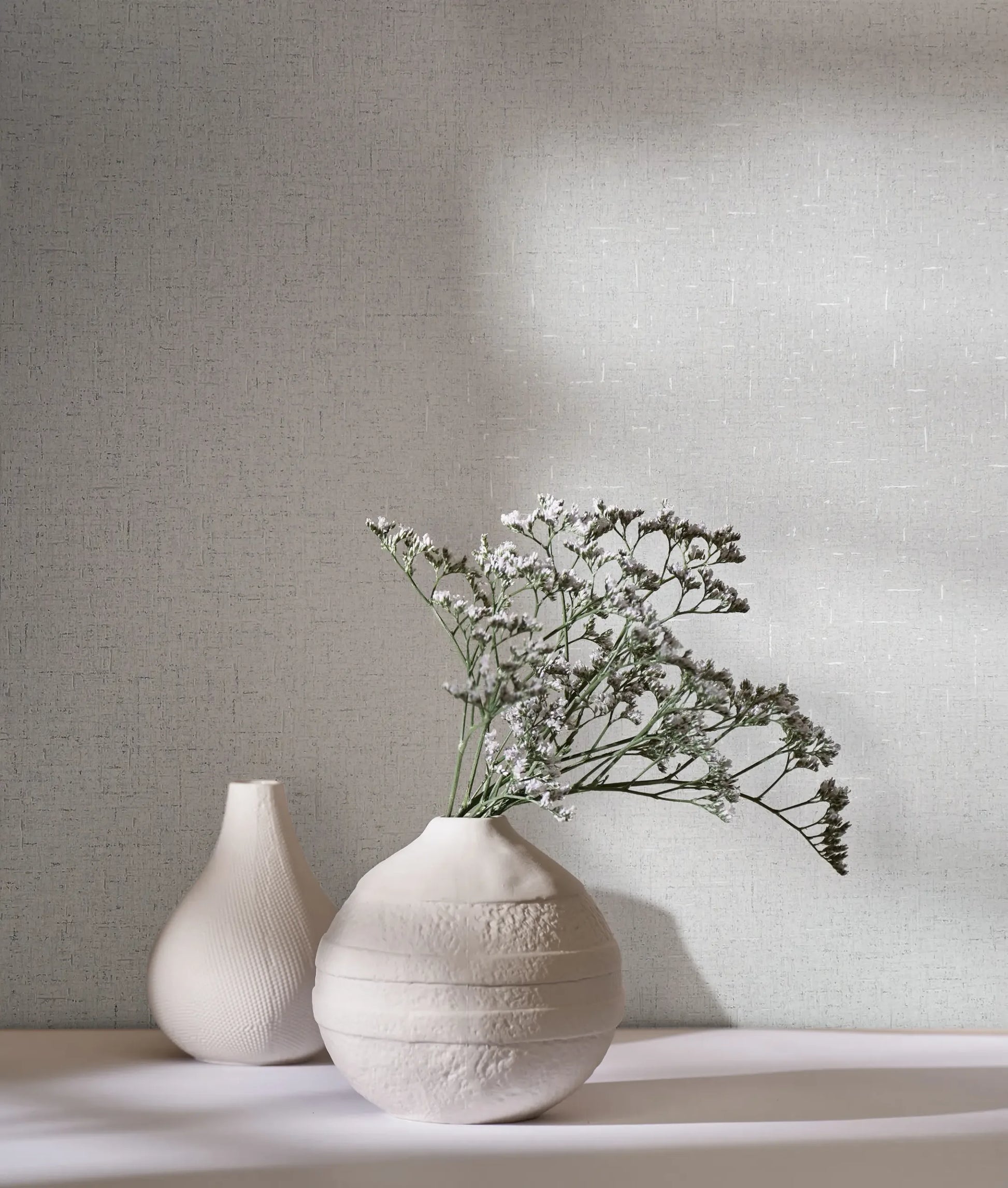 Marburg City Romance 35109 Light gray crosshatched wallpaper behind two white ceramic vases, one holding small white flowers, with soft shadows across the scene.