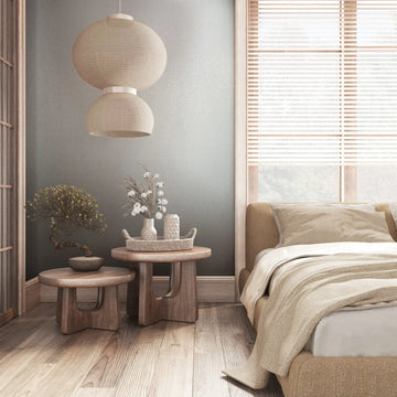 Marburg City Romance 35109 Gray textured wallpaper behind a cozy bedroom scene with a bed, two round wooden side tables holding a potted plant and vases on a woven tray, a pendant light, and a window with blinds.
