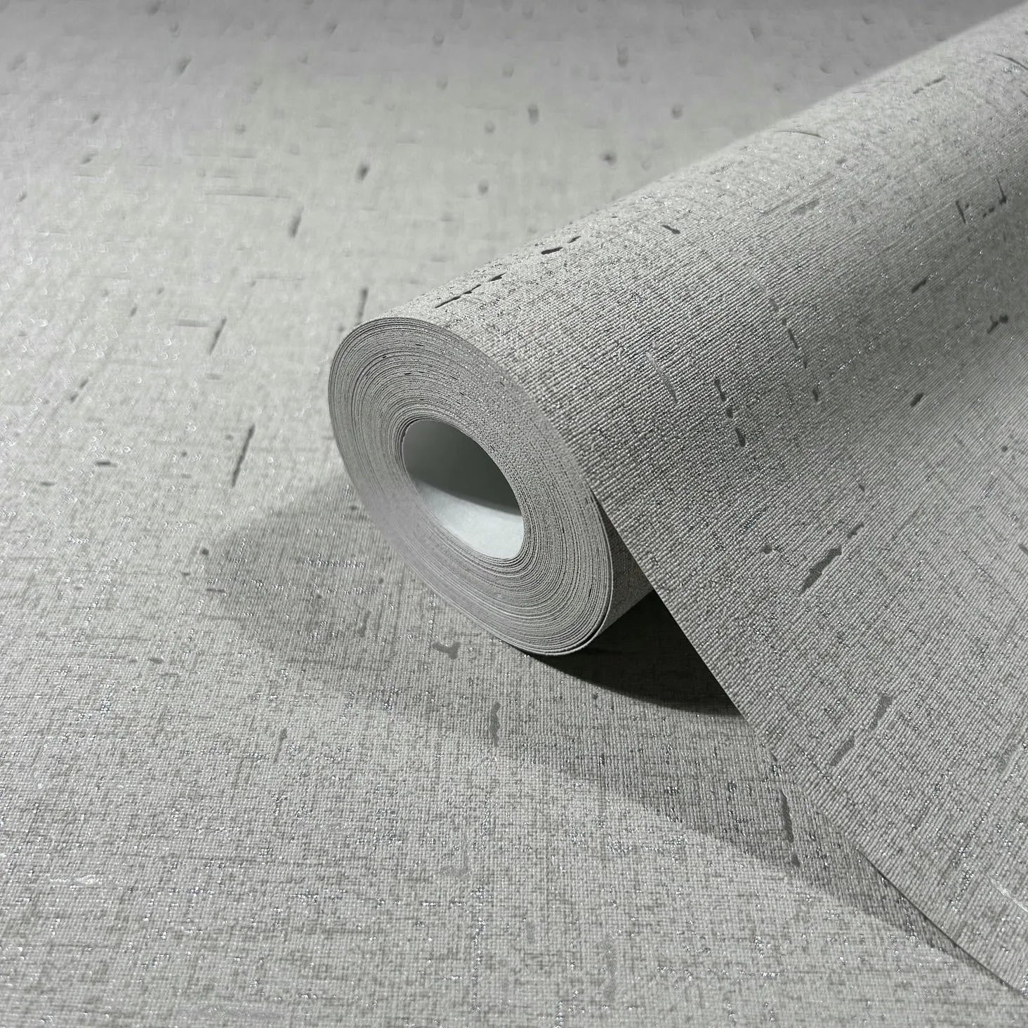 Marburg City Romance 35110 Close-up of a gray textured wallpaper roll resting on a matching unrolled sheet, showing a woven-look surface with scattered short dash marks and a subtle shimmer.