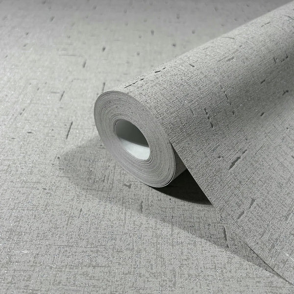 Marburg City Romance 35110 Close-up of a gray textured wallpaper roll resting on a matching unrolled sheet, showing a woven-look surface with scattered short dash marks and a subtle shimmer.