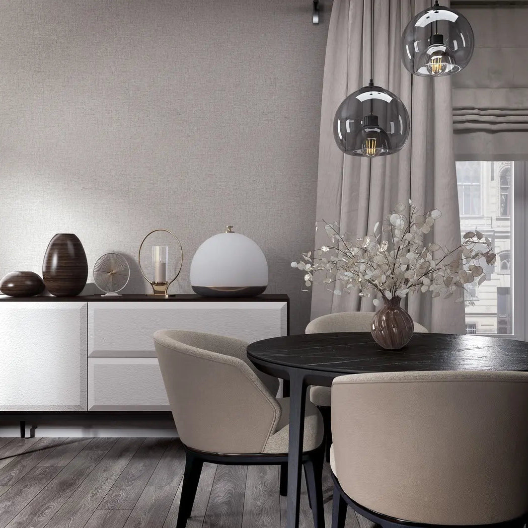 Marburg City Romance 35110 Light gray textured wallpaper with a subtle linen look behind a modern dining area featuring a round black table, beige chairs, a white sideboard with decor, glass pendant lights, and a window with drapes.