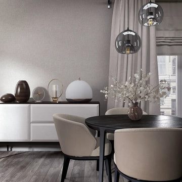 Marburg City Romance 35110 Light gray textured wallpaper with a subtle linen look behind a modern dining area featuring a round black table, beige chairs, a white sideboard with decor, glass pendant lights, and a window with drapes.