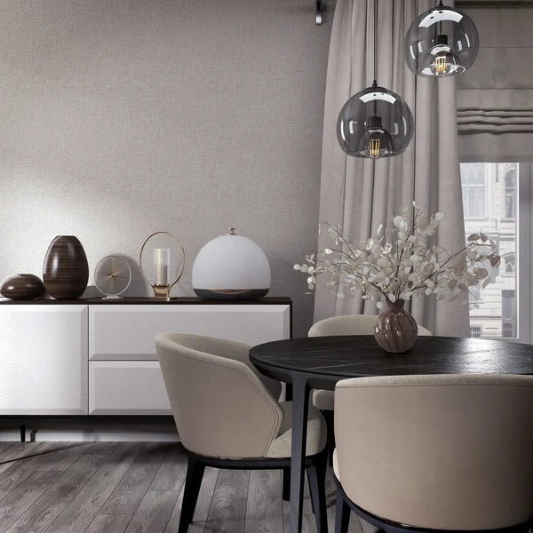 Marburg City Romance 35110 Light gray textured wallpaper with a subtle linen look behind a modern dining area featuring a round black table, beige chairs, a white sideboard with decor, glass pendant lights, and a window with drapes.