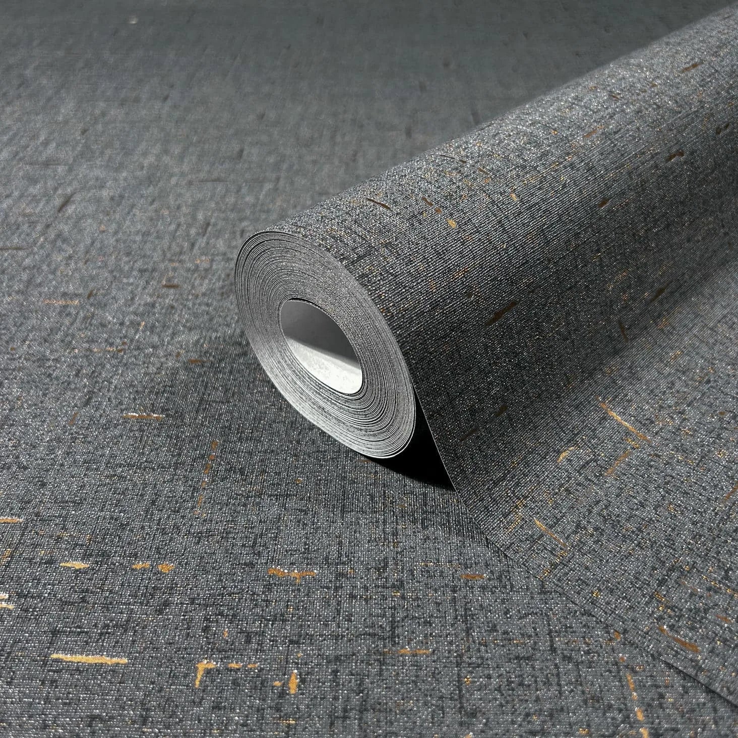 Marburg City Romance 35111 Close-up of a dark grey wallpaper roll with a fine crosshatch texture and scattered gold flecks, resting on the same pattern.