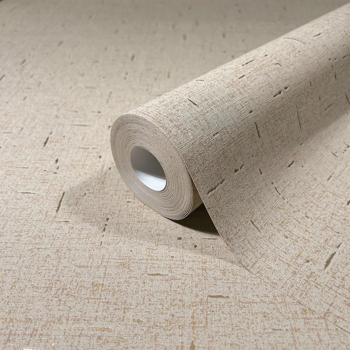Marburg City Romance 35112 Close-up of a beige wallpaper roll on matching unrolled wallpaper, showing a fine linen-like texture with scattered short linear flecks.