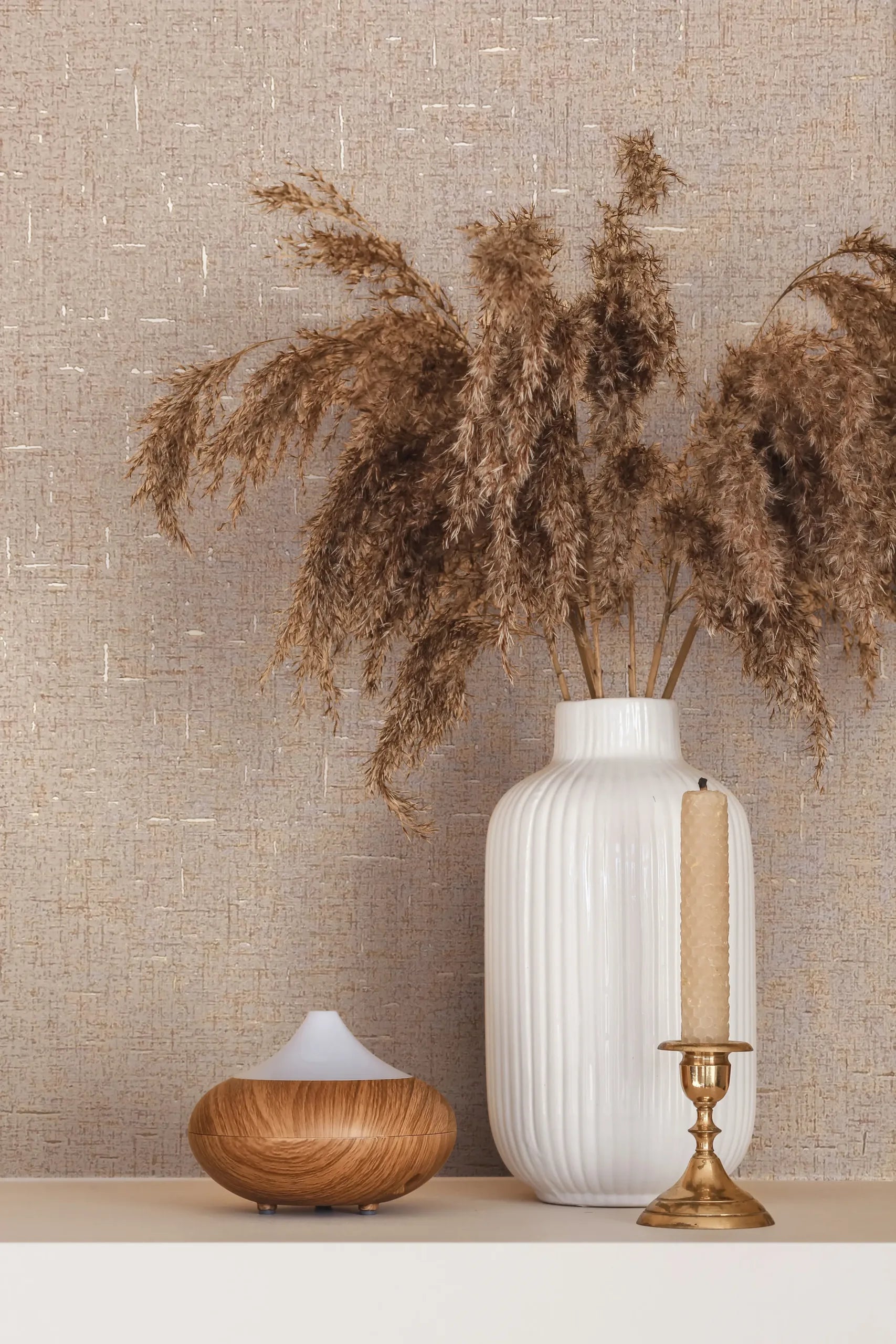 Marburg City Romance 35112 Beige crosshatched wallpaper behind a white ribbed vase filled with dried pampas, a brass candlestick with a taper, and a small wood‑grain diffuser on a light shelf.
