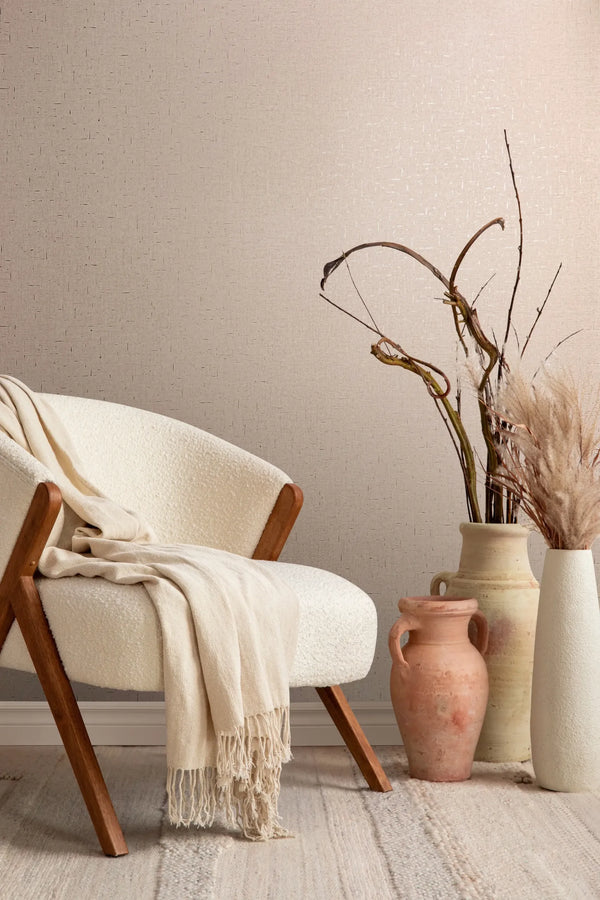 Marburg City Romance 35112 Beige wallpaper with a subtle crosshatch dash texture behind a cream armchair with a draped throw and several ceramic vases holding dried branches and pampas on a light rug.