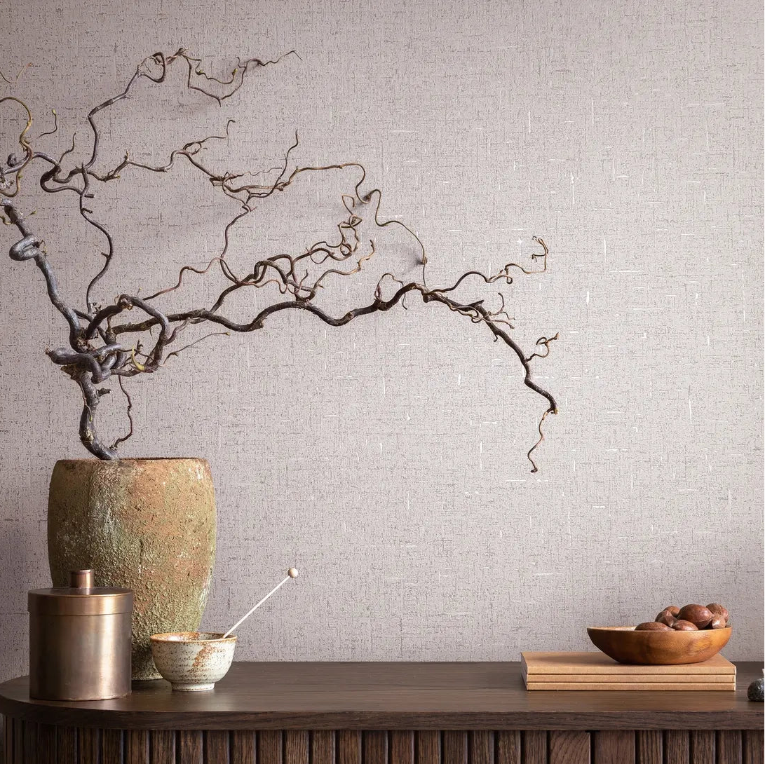 Marburg City Romance 35113 Beige-gray textured wallpaper with a fine crosshatch pattern behind a console holding a tall ceramic vase with twisted branches, a metal canister, a small bowl with a spoon, stacked books, and a wooden bowl of nuts.