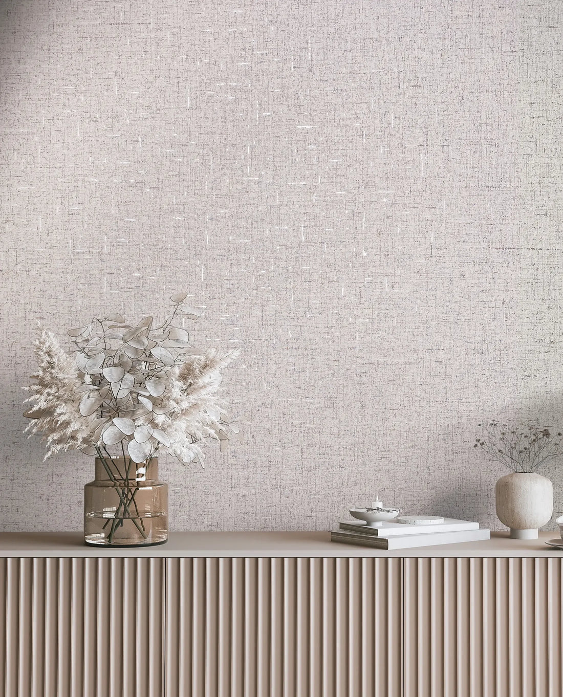 Marburg City Romance 35113 Beige-gray textured wallpaper with a fine crosshatch pattern behind a console holding a tall ceramic vase with twisted branches, a metal canister, a small bowl with a spoon, stacked books, and a wooden bowl of nuts.