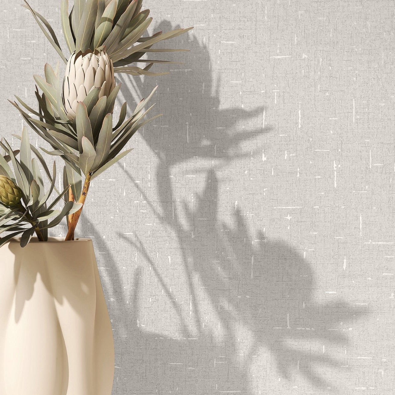 Marburg City Romance 35114 Beige textured wallpaper with small white dashes, shown behind a cream vase holding leafy stems with strong plant shadows on the wall.