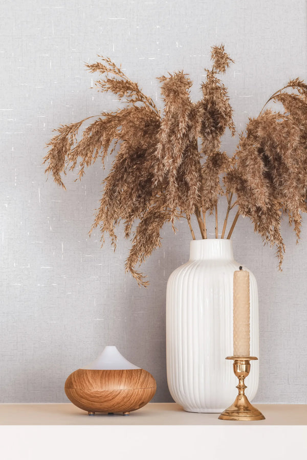 Marburg City Romance 35115 Close-up styled scene showing pale textured wallpaper with a fine crosshatch and small white slubs behind a ribbed white vase of dried pampas, a brass candlestick with a beige taper, and a wood‑grain diffuser on a shelf.